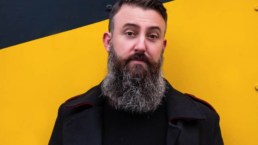 A man with big busy beard standing against bright yellow and black wall.