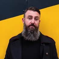 A man with big busy beard standing against bright yellow and black wall.