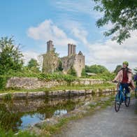 Two cyclists on a path by a river with the ruins of a castle in the background