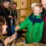 A adult and child touching the pointed end of an arrow head being help up by a woman seated at a table with other ancient artifacts.