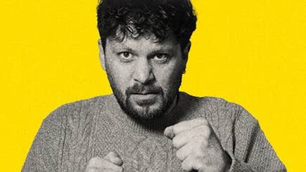 A man looking a bit fierce holding up his hands in fists in front of him, against bright yellow background.