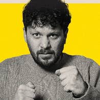 A man looking a bit fierce holding up his hands in fists in front of him, against bright yellow background.