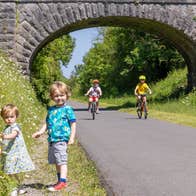 Two kids on bikes along the Old Rail Trail Greenway along with two kids that are picking flowers on the side of the trail