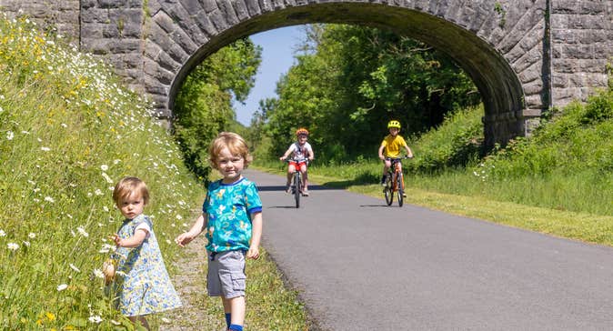 Two kids on bikes along the Old Rail Trail Greenway along with two kids that are picking flowers on the side of the trail
