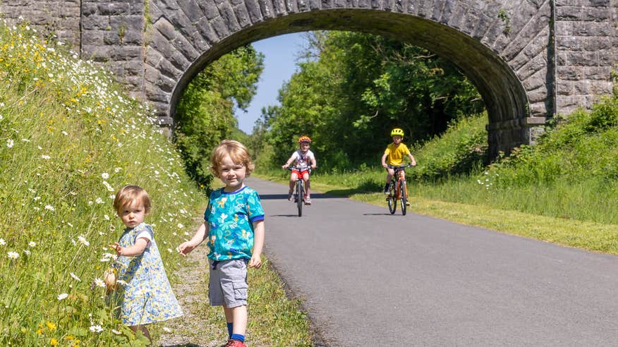 Two kids on bikes along the Old Rail Trail Greenway along with two kids that are picking flowers on the side of the trail