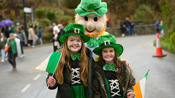 2 young girls dressed in green outfits posing with a person dressed as a leprechaun outdoors on a road.