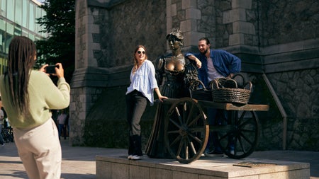 A group of friends posing with the Molly Malone statue while another friend takes their picture.