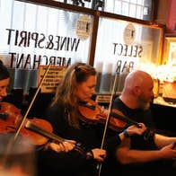Three musicians sitting in a pub playing instruments with a window behind them