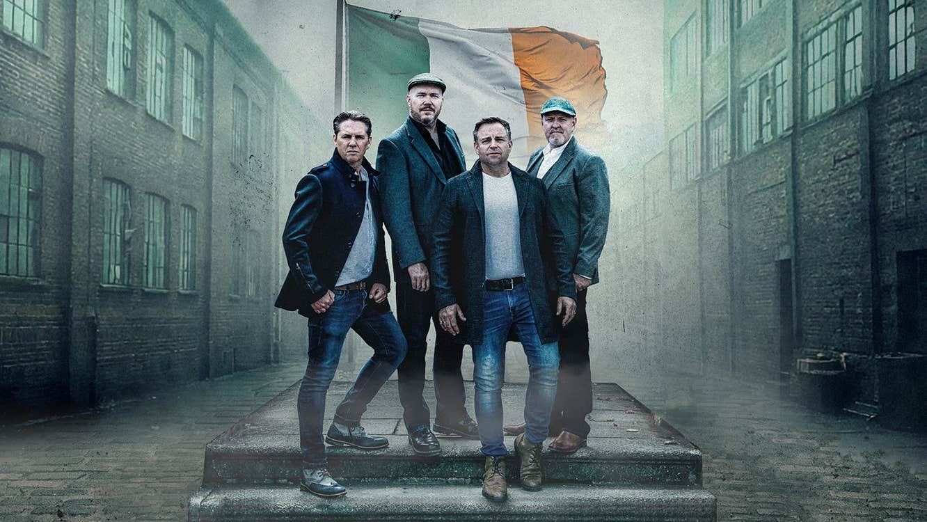 4 serious men in a group on 2 steps standing in front of Irish flag with old industrial buildings either side
