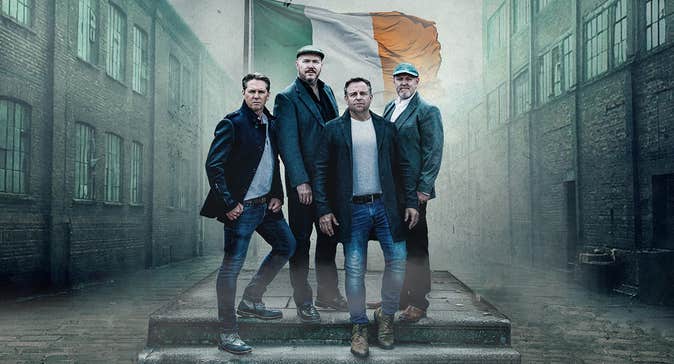 4 serious men in a group on 2 steps standing in front of Irish flag with old industrial buildings either side