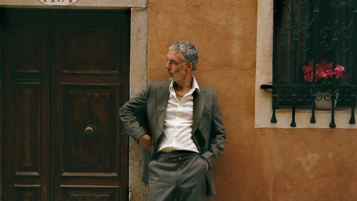A man in a casual suit leaning against a brown building beside a dark wooden door