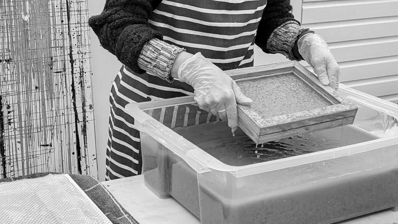 A pair of hands lifting a small wooden square frame over a large plastic container holding a dark liquid.