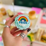 Pride Month - Afternoon Tea Trip - a hand holding out a small cup cake with swirl of icing on and a small rainbow logo, against blurry background of tea setting