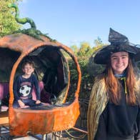 A smiling girl in black witches hat and costume with orange pumpkin style small carriage behind with young boy seated in it.