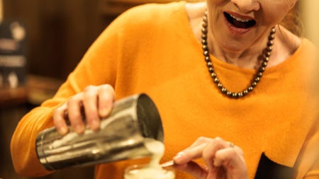 A woman pouring cream onto a brown liquid in a cocktail glass