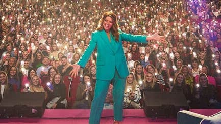Doireann Garrihy - The Laughs Of Your Life Live, a smiling woman with arms outstretched on a stage with large audience in the background holding lights.