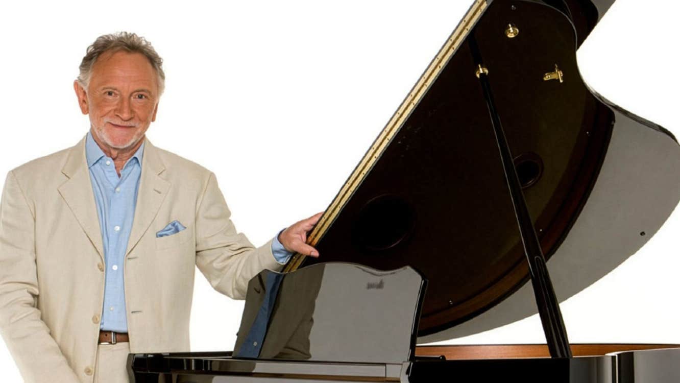 Phil Coulter in Concert Dec 2026, a smiling man in light coloured suit standing beside a grand piano with the lid open.