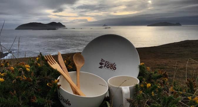 A white bowl, plate and mug holding set of wooden cutlery set against sea and rocky background.
