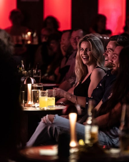 Audience members sitting at at table with drinks and laughing with red lights in the background
