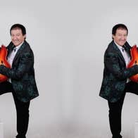 A smiling man in dark suit with one foot up on small white block, holding up a guitar vertically