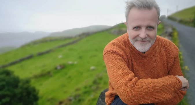 A smiling man with crossed arms outdoors with background of green hills