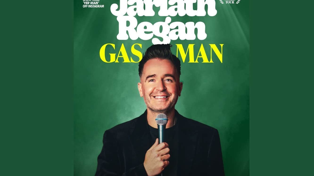 Man dressed in black, smiling holding a mic up to his face, against dark green background.