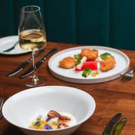 Table with two white plates of food a glass of wine and cutlery on it