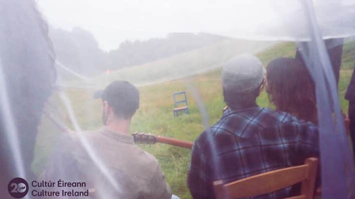 rear view of 3 people seated on wooden chairs in a grassy field, viewed through white net curtain.