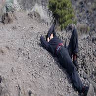 still of a woman dressed in all black lying on the slopes of Mount Vesuvius. Her right hand is behind her hand is behind her head and she raises her left arm over her head.