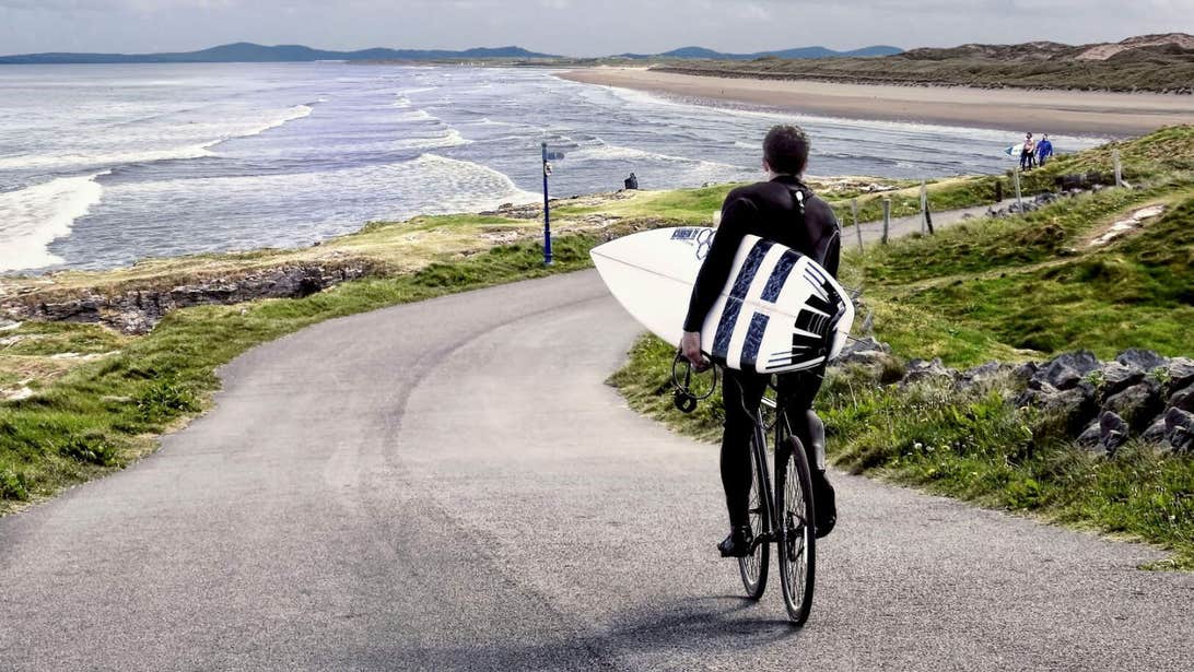Surfer holding his surfboard, cycling down toward Tullan Strand in Bundoran.