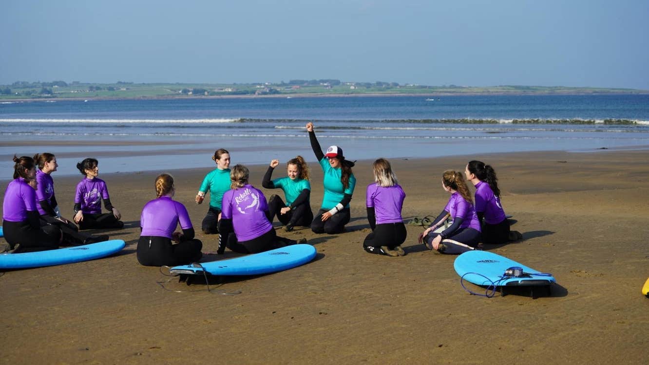 Group of surf students with surfboards sitting in a circle on the beach during a lesson
