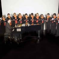 Festival of Voice: Cór Ban Cúil Aodha, a group of women standing in a semi circle around a small grand piano being played by a man.