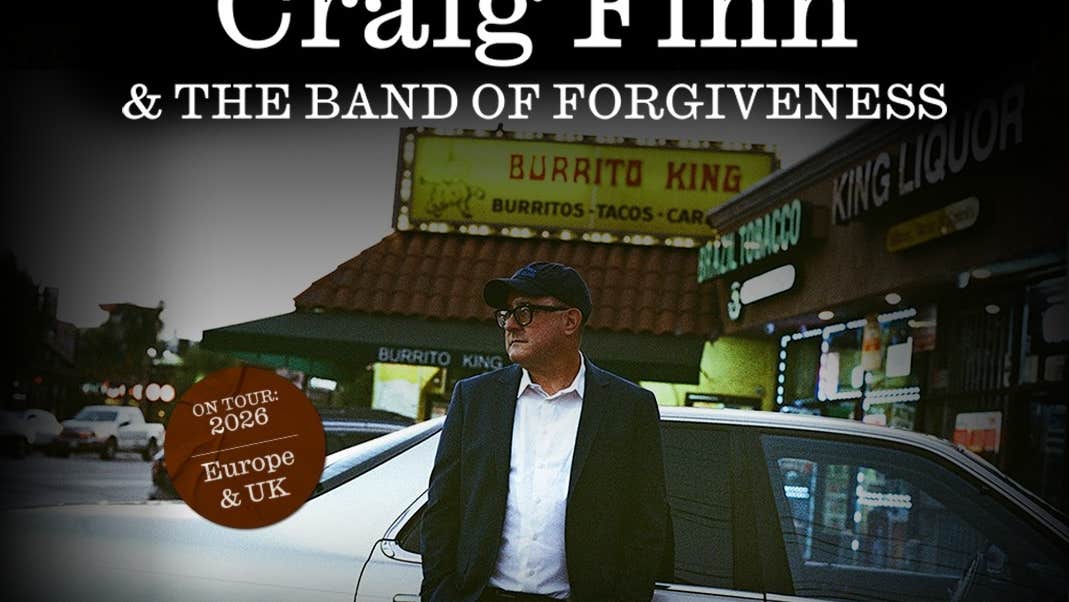 A man in jacket, white shirt and peaked cap is leaning against a silver car outside shops