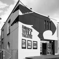 Black and white image of exterior of a theatre with three posters on the wall and railings to the left