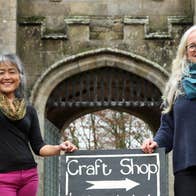 Two ladies standing outside a castle like gate lodge beside a sign that says crafts with an arrow pointing to the right