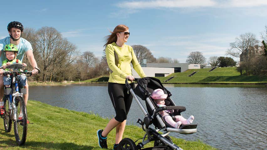 Man on a bike with a child while a woman pushes a child on a buggy beside a river