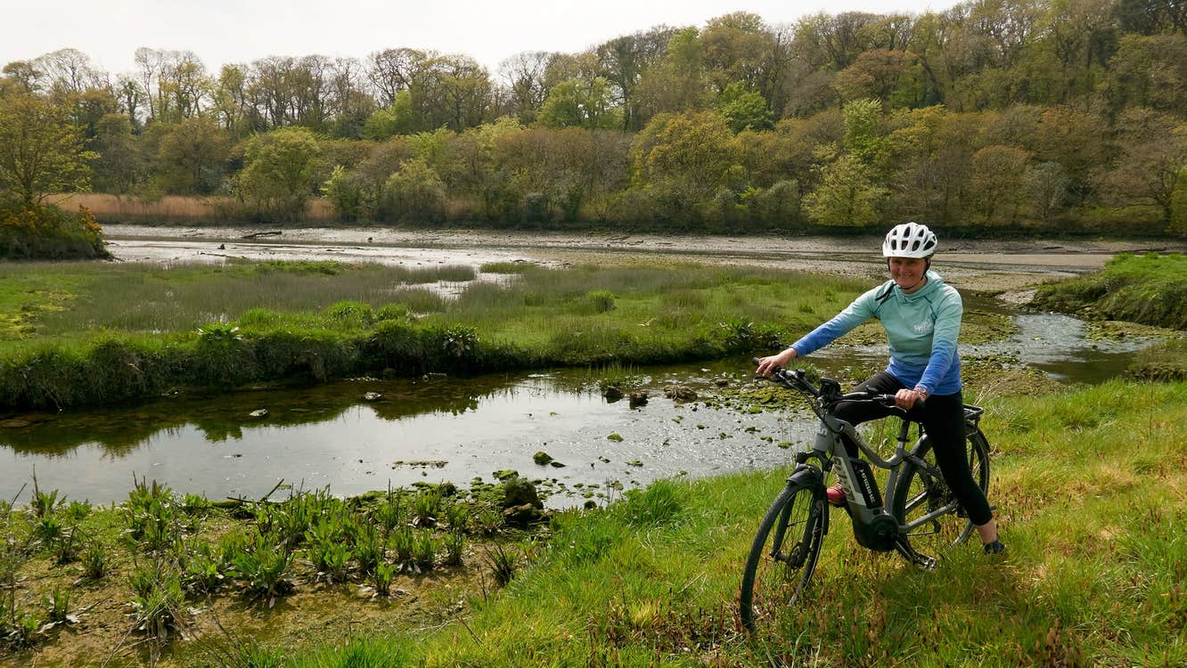 Person sitting on a bike in a field next to a river with trees in the background