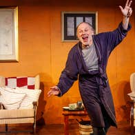 On a stage with armchairs, a man in a dressing gown is waving his arms looking excited.