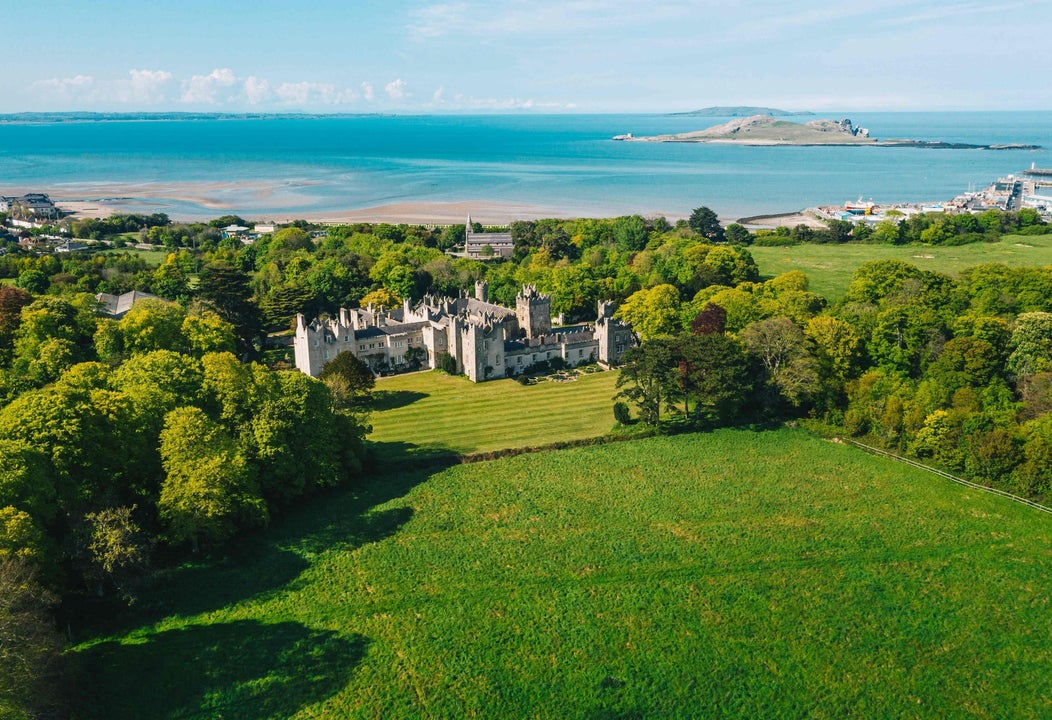 Aerial view of a castle surrounded by green fields and trees and a beach in the background