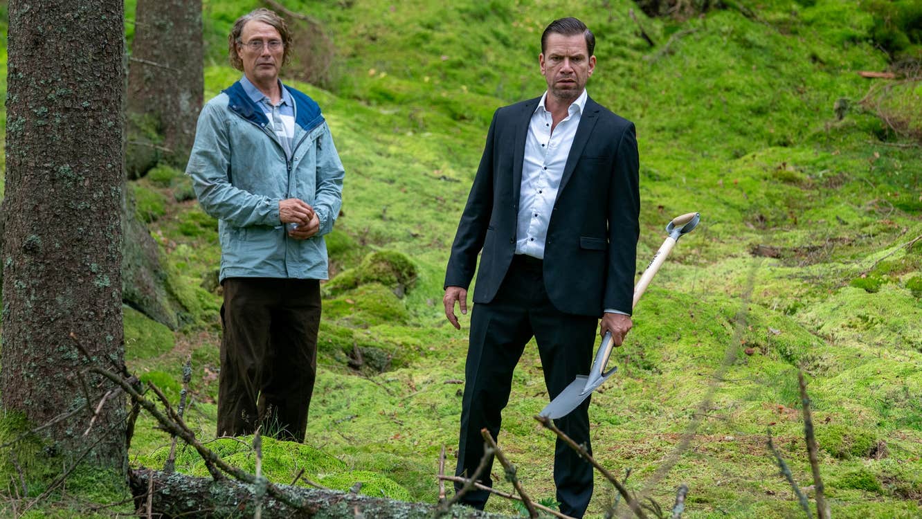 A man in a crumpled raincoat and a man in a suit are in a forest.