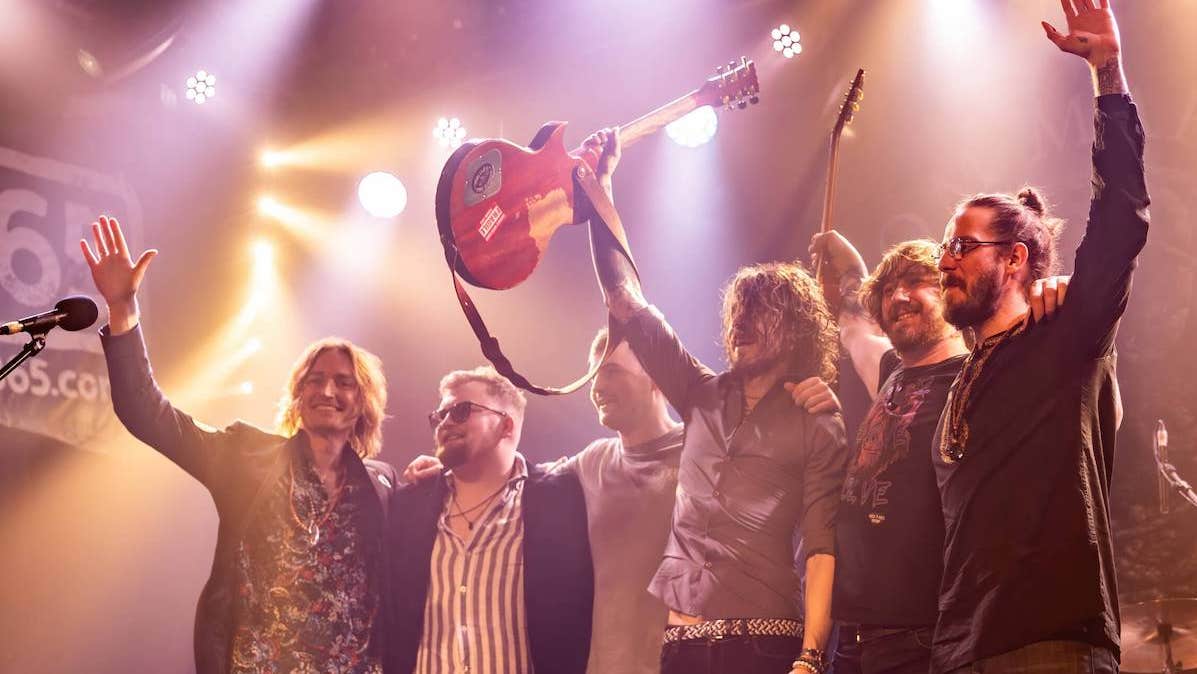 6 men in a line with arms around each other at the front of a stage, some holding up their arms and smiling.