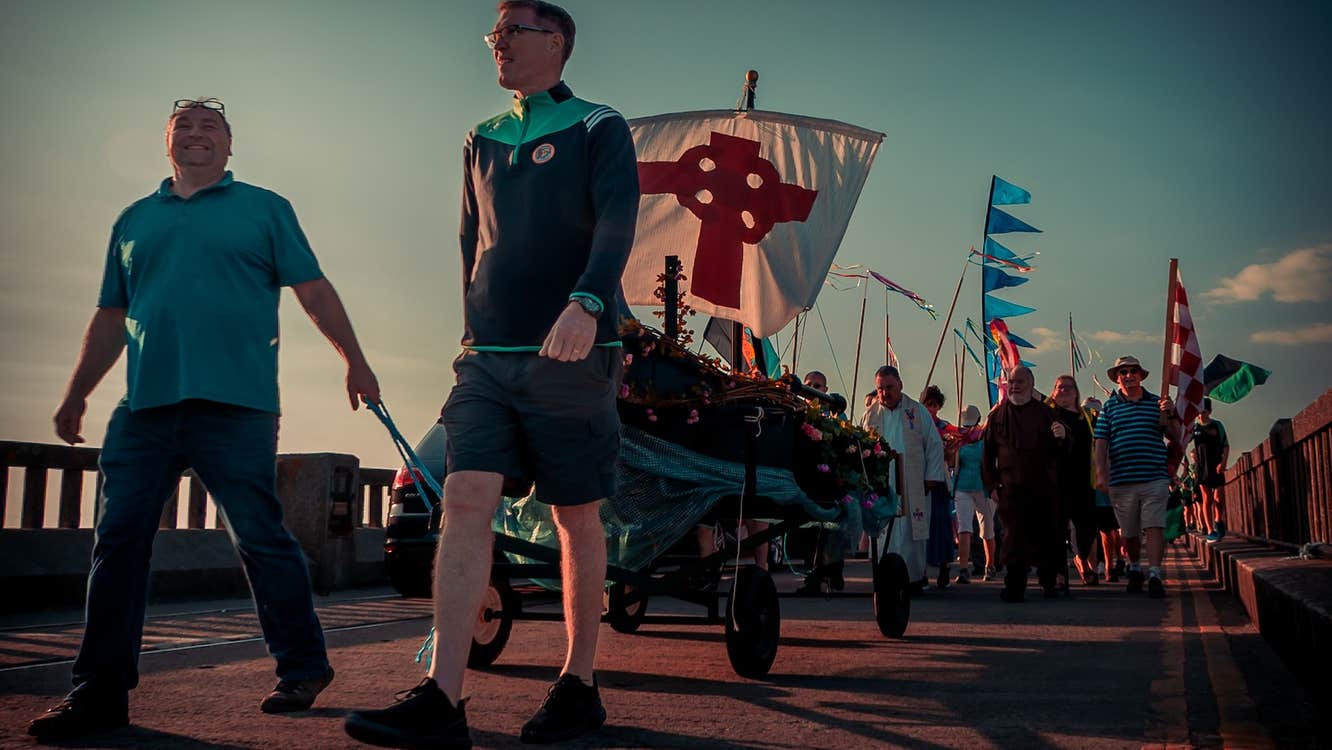 St Brendan the Navigator Community Festival 2026, at dusk, 2 men are pulling a small trolley in a parade with flags.