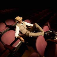 A man in waistcoat and shirt is seated in red cinema type seats with his feet up on the back on the seat in front, in darkness illuminated by a spot light.