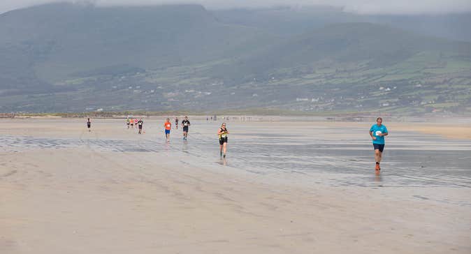 View of a large beach with people running along it, against background of hills