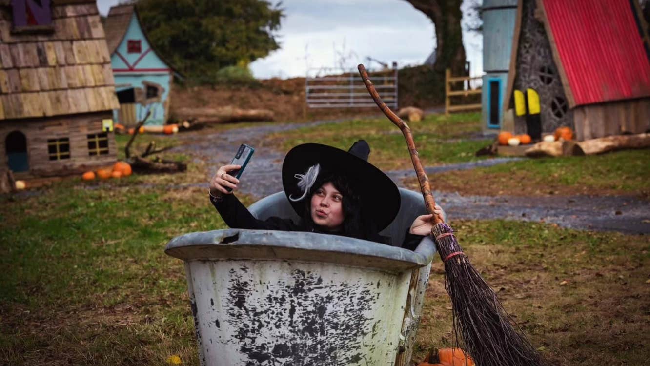 A person with large black hat and broom is in a large grey container taking a selfie.