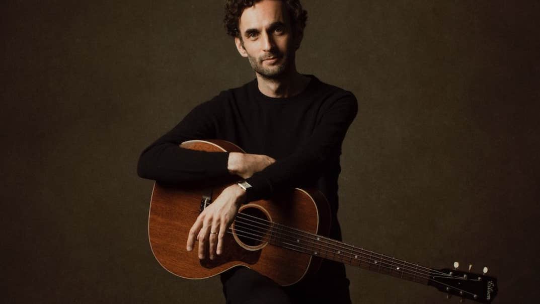 A man in black clothes sitting on a stool, resting his arms on a brown guitar.
