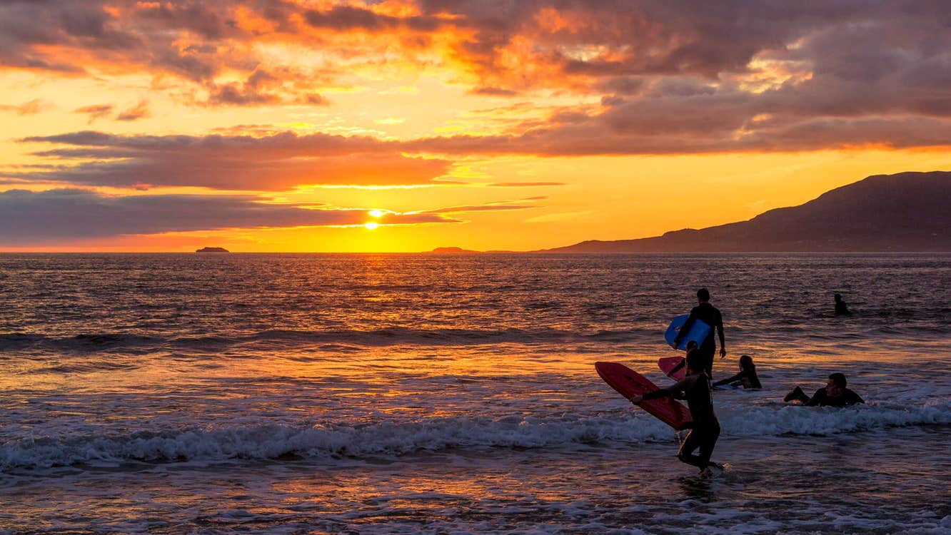 People running into the water at Carrowniskey Strand with surfboards at sunset.
