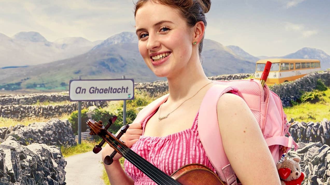 Ciara Cox holding a violin standing in front of a Gaeltacht sign and bus with stone walls and mountains in the background