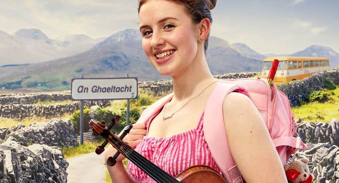 Ciara Cox holding a violin standing in front of a Gaeltacht sign and bus with stone walls and mountains in the background