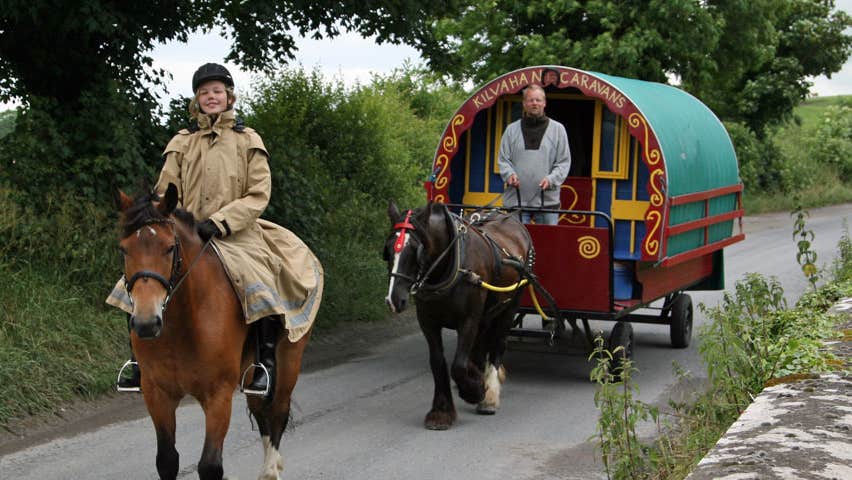 A horse drawn carriage riding along on the road with a woman on another horse riding in front